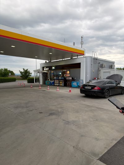 A black Mercedes-Benz with license plate PA3211KP is parked at a Shell gas station convenience store labeled "SELECT" in Dragor, Bulgaria. The hood of the car is open. Orange and white traffic cones delineate a path along the gas station's concrete apron. A person is holding a cell phone in the upper right corner of the image. The sky is overcast.