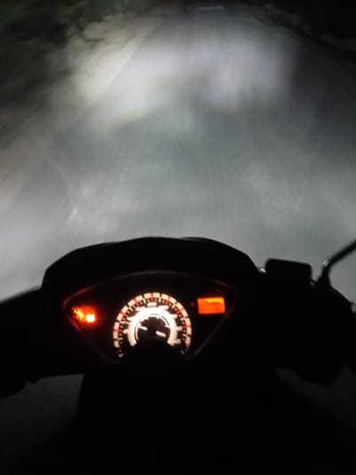 This image shows a first-person perspective of riding a motorcycle at night. The headlight illuminates a foggy or misty road ahead. The motorcycle's dashboard is visible in the foreground, with a speedometer showing a speed of approximately 40 km/h. A turn signal indicator is lit on the left side of the dashboard. The background is dark and blurred, with the light from the headlight creating a hazy effect on the road and surrounding foliage. The specific location in Cotkumbang, Indonesia, is not discernible from the visual information presented.