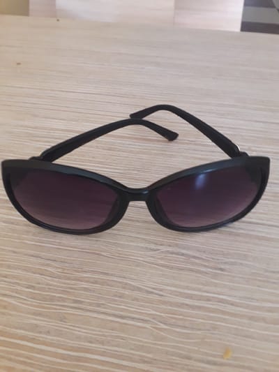 The image displays a pair of black sunglasses resting on a light-colored wooden surface. The photograph is oriented vertically, causing the horizontal grain of the wood to appear as vertical lines across the frame. The sunglasses are the central subject, featuring dark, possibly purple-tinted lenses and a classic, rounded frame. One arm of the sunglasses is folded inward, while the other is extended.

The wooden surface, likely a table or floor, shows a fine, textured grain pattern. In the upper left corner of the original image, and partially visible in some crops, there is a hint of another background element, possibly a wall or furniture, characterized by subtle vertical stripes in muted shades of grey, white, and light brown. A tiny, faint yellow speck is visible on the wooden surface towards the right side of the frame in some detailed crops.

No activity is taking place; the sunglasses are stationary. The lighting is even and soft, indicative of an indoor environment. There are no people visible, and consequently, no emotions are conveyed. No text is visible on the sunglasses or within the background. The scene depicts an inanimate object in an indoor setting in Ghafurov, Tajikistan.
