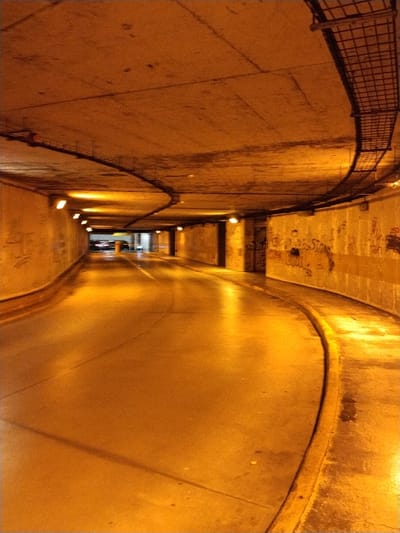 A curving tunnel with bare concrete walls and ceiling is illuminated by warm, yellow lighting. Graffiti marks the right wall. A metal cable tray runs along the upper right curvature of the tunnel. The floor is smooth and reflective, casting light upwards. In the distance, at the tunnel's exit, dark vehicles are faintly visible. The scene is dominated by the amber hue of the lights.