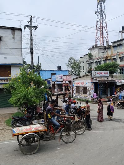 The image depicts a street scene in Benapole, Bangladesh, on an overcast day. In the foreground, a three-wheeled bicycle cart, loaded with what appear to be sacks or bags, is parked on the asphalt road. A man in a dark t-shirt and jeans is seated on the cart, holding onto the handlebars. To the left of the cart, a black motorcycle is parked.

Further back, several individuals are gathered. A woman in a floral patterned dress and headscarf stands facing right, holding a phone. Beside her, a woman in a purple and black patterned outfit, with a light-colored headscarf, stands with a bundle in her arms. A man in a striped shirt and black pants stands near them.

The background reveals a mix of commercial establishments and residential buildings. One building prominently displays signage with Bengali script, including "শাঁখা দই" which translates to "Shakha Doi" (a type of yogurt). Another building has a large telecommunications tower with red and silver lattice work on its roof. Electrical poles with numerous wires are visible throughout the scene. Trees with green foliage and yellow flowers add a touch of nature to the urban environment. The overall impression is that of a bustling local market or roadside area.