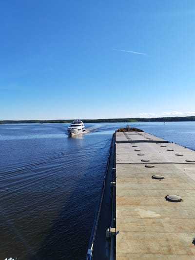 This image captures a wide-angle view of a large waterway under a clear blue sky, identified as being near Novodevich'e, Russia. In the foreground on the right, the weathered, flat deck of a large, rust-stained barge or cargo vessel occupies approximately two-thirds of the lower right frame. This deck features several evenly spaced, circular metal hatch covers. The left side of the barge's deck shows a dark railing and some associated hardware. To the left of the barge, dark blue water exhibits ripples and small waves.

In the midground, a white, multi-deck motor yacht with a modern design is underway, approaching the viewer from the center-left. The yacht is actively creating a V-shaped wake behind it, indicating forward motion. Its bow is pointed towards the viewer, and reflections are visible on its hull.

The background features a continuous, dark green treeline or forest stretching across the horizon, suggesting distant land on either side of the expansive body of water. The sky above is a uniform, vibrant blue with minimal cloud cover, though a few faint, wispy cirrus clouds are discernible in the upper portions of the sky. No individuals are visibly present on either the yacht or the barge.