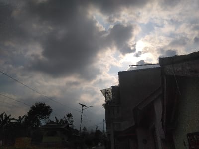 The image captures a semi-urban street scene in Houayyen, Laos, dominated by a heavily clouded sky. Dense, dark grey cumulus clouds occupy the majority of the sky, with bright patches of sun visible in the upper right quadrant, creating a strong backlight and glare. Utility lines run horizontally across the upper left and mid-left areas. On the left, a cluster of dark green trees and foliage partially obscures a small, light-roofed building. A black lamppost with a solar panel mounted on its top stands in the midground. The right side of the frame is occupied by the dark, silhouetted facades of several multi-story buildings, appearing to have rough, possibly concrete, textures. One building exhibits a partial metal or mesh structure on its upper section. No individuals or discernible actions are present.