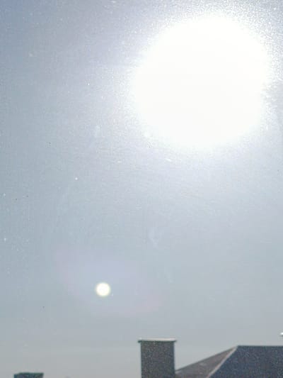 The image shows a bright, hazy sky with the sun prominently displayed in the upper right quadrant. The sun's glare is intense, causing overexposure in that area. Below the bright glare, a smaller, distinct circular shape, possibly another reflection or lens flare, is visible in the upper center of the frame. The sky appears overcast or hazy, with a diffused light that obscures finer details.

In the lower portion of the image, the rooftops of buildings are visible. These appear to be industrial or residential structures, with dark, angular roofs and chimney-like elements. The overall scene suggests an outdoor setting, likely a view from a window or elevated position looking out towards a sky and cityscape. The lighting and atmospheric conditions indicate it is daytime, with the sun high in the sky, but the haziness suggests a potential for diffused sunlight, possibly due to clouds, fog, or pollution. No people are visible in the image, and there is no visible text. The location context of Smarhon, Belarus, is provided, suggesting this is a view of that region.