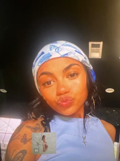 A young woman is captured in a selfie, posing with her lips puckered. She has dark, wavy hair and is wearing a blue and white patterned headscarf. Her outfit consists of a light blue tank top, adorned with a delicate silver cross necklace. Her left arm is visible, showcasing a butterfly tattoo. In the lower portion of the image, a small thumbnail displays a person in a white dress. The background is dark, with a rectangular vent visible on the upper right. The overall lighting suggests an indoor setting. There is no visible indication of the location being in the city of Boone, United States of America.