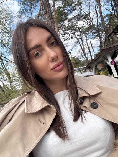 A young woman with fair skin and long brown hair poses for a selfie in Konstancin-Jeziorna, Poland. She has light eyes and is wearing a beige trench coat over a white top. The photo is taken outdoors, with trees and a partially visible white building or structure in the background. The sky is visible through the branches.