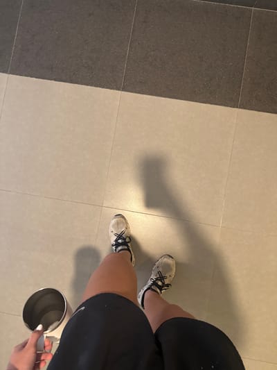 An overhead shot captures the lower body of an individual standing on a tiled floor. The person wears black athletic shorts, revealing tanned legs, and white athletic shoes with black laces and dark soles, which appear somewhat scuffed. The left hand, with white-painted fingernails, holds a silver-colored, cylindrical cup with a white handle. The floor is composed of light beige tiles in the foreground and a darker, speckled gray-black tile section in the background, meeting at a diagonal line across the frame. Shadows of the person's legs and the cup are visible on the light-colored tiles, along with a larger shadow suggesting the presence of the upper body. The image does not contain visual information to confirm a location within Santa Catarina, Mexico.
