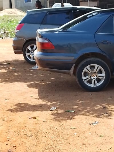 The image depicts an outdoor scene in Oke Onigbin, Nigeria, characterized by an unpaved reddish-brown dirt surface. Two passenger vehicles are parked side-by-side, facing away from the camera. In the foreground, a dark blue sedan, likely a Toyota Corolla model, is positioned with its right rear quarter visible. It features a prominent taillight assembly with red and clear sections, a dark lower bumper trim, and a multi-spoke silver alloy wheel on the right rear axle. Immediately behind this sedan, a grey or dark green station wagon or hatchback is partially visible, showing its left rear quarter and a similar multi-spoke silver alloy wheel.
A single individual, a dark-skinned person wearing a dark top, is partially observed standing behind the grey vehicle, looking towards the right of the frame. The background includes a light-colored building with a blue window and what appears to be a pile of construction material or dirt. The ground shows scattered debris, including pieces of paper and plastic, along with shadows from an unseen overhead source, indicating daylight conditions. No overt actions or interactions are discernible.