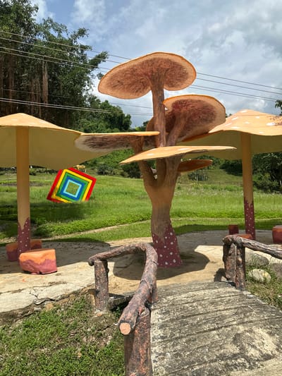 The image shows a decorative outdoor area with mushroom-themed sculptures. A central mushroom sculpture features a thick, textured trunk with multiple caps of varying sizes stacked atop each other, resembling a cluster. The caps are a light orange-brown color with darker undersides that show radiating lines. The base of the central mushroom and the stems of surrounding mushroom structures are decorated with red and white spots.
To the left and right of the central sculpture are two larger mushroom structures with wide, flat caps. The cap on the left is supported by a thick, spotted stem, and a colorful, square mosaic featuring concentric squares of red, yellow, green, blue, and white hangs from beneath it. The cap on the right is also supported by a spotted stem.
In the foreground, there is a small, rustic-looking bridge made of what appears to be faux wood material, with a textured surface. The bridge has railings made of the same material, designed to resemble tree branches. Two cube-shaped, multi-colored seating elements, red and orange, are placed on the ground near the base of the mushroom structures.
The environment is grassy and appears to be a park or garden. In the background, there are trees, a slight incline of land, and power lines running horizontally across the upper portion of the image. The sky is partly cloudy. The overall scene suggests a whimsical or fantasy-themed recreational space. The location is identified as Khánh Vĩnh, Vietnam.