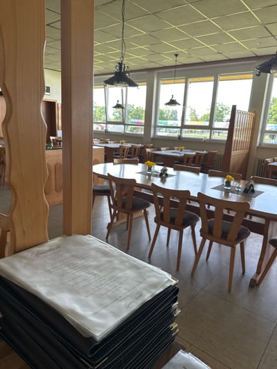 The image shows an interior view of a restaurant. There are rows of wooden tables and chairs leading into the restaurant, and daylight streams in through large windows. A stack of menus is in the foreground, indicating that the establishment is open and ready to serve customers. A menu leans against a wooden chair and displays the German word "Speisekarte" indicating the menu of the establishment. The OCR text on the menu includes the word "Herxheim" and prices in Euros, indicating that the location is in the city of Herxheim, Germany. The menu lists various food items, including "Rumpsteak" and "Schnitzel."