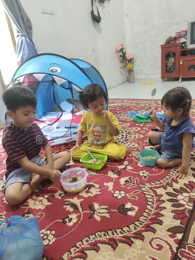 The image depicts three young children sitting on a red oriental rug indoors, possibly in a residential area of Cintadamai, Indonesia. A blue shark-shaped play tent is visible behind them. The child on the left, a boy wearing a striped shirt and shorts, is looking down and appears to be eating from a plastic container. In the center, a girl in a yellow t-shirt and pants is also eating from a green bowl. To the right, another girl in a blue denim outfit is holding a spoon and looking towards the center. Several bowls and a blue plastic bag are scattered around the children. The environment suggests a casual, domestic setting, with a plain wall and some furniture visible in the background.