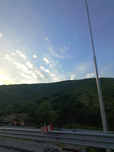 The image depicts a wide landscape scene, likely captured during daytime with the sun low on the horizon, casting a warm light on the clouds and sky. The scene is dominated by a densely forested, dark green hillside that occupies the middle and lower sections of the frame. A clear blue sky with scattered white clouds stretches across the upper portion. A tall, metallic pole, possibly a street light or utility pole, stands vertically on the right side, extending from the bottom to near the top of the image. In the lower foreground, a section of a road barrier with horizontal silver rails is visible, with a red and white striped marker pole positioned in front of it. Beyond the road barrier, to the left, a few buildings with reddish-brown roofs are nestled on the lower slope of the hillside. Several cars are parked or driving in front of these buildings, suggesting a small settlement. The overall impression is that of a rural or semi-rural setting, possibly near the outskirts of a town or village, with nature playing a significant role in the scenery. The location is identified as being in the city of Lyutidol, Bulgaria.