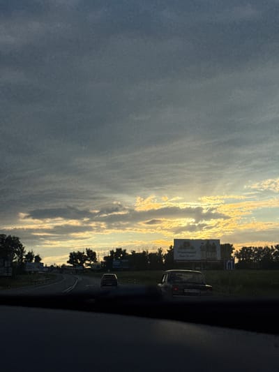 The media file captures a scenic view from a car traveling on a road, likely in Novomar'evskaya, Russia, during sunset. The sun, low on the horizon, casts a warm golden glow through the clouds, creating a dramatic and serene atmosphere. The sky is a mix of dark gray and bright yellow, with streaks of light illuminating the scene.
On the right side of the frame, trees line the road, their silhouettes stark against the vibrant sky. A dark-colored car is visible on the road ahead, its taillights illuminated. In the background, a billboard with text is partially visible, along with other signs and structures suggesting a roadside environment. The interior of the car, from which the photo is taken, is dark and out of focus, emphasizing the view outside. The overall mood is peaceful and contemplative, possibly evoking a sense of journey or transition as the day ends.