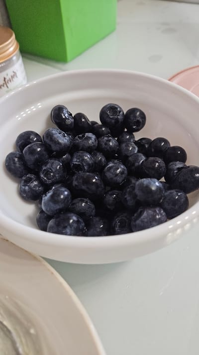 The image shows a white bowl filled with blueberries. The bowl is partially visible, with some of the blueberries spilling slightly over the edge. The blueberries appear fresh and are a deep blue color. In the background, there's a green rectangular object, a tan colored jar, and a white surface. There is no way to know if this image was taken in Yerzovka, Russia or anywhere else for that matter. There are no people, actions, or interactions visible in the image.