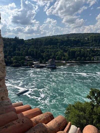 The image depicts a scenic view of the Rhine Falls in Dachsen, Switzerland. The primary subjects are the powerful, turbulent water of the falls, which is a vibrant turquoise color with white frothy waves. A few boats are visible on the water, one with people on board, suggesting a tourist activity. The river is bordered by a lush, green, densely forested hill that slopes down to the water's edge. A few buildings, likely restaurants or visitor facilities, are situated along the riverbank, nestled among the trees. In the foreground, the edge of a stone wall and some reddish-brown, cylindrical objects, possibly roof tiles or decorative elements, frame the view.
The scene is set under a bright sky filled with dramatic, fluffy white clouds against a clear blue backdrop, indicating fair weather. The lighting suggests it is daytime, likely late morning or early afternoon, with sunlight glinting off the water. The overall mood is one of natural grandeur and outdoor leisure. The image is captured from an elevated vantage point, looking down and across the falls. No visible text is present in the image.