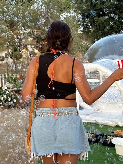 The image captures a rear view of a woman standing outdoors, surrounded by numerous floating bubbles. She is wearing a black crop top with a cutout detail and neon coral straps, paired with a light blue denim skirt. A woven bag with a leather strap is slung over her right shoulder. A decorative gold belt with star-shaped accents cinches her waist. In the background, a white inflatable structure is visible, and beyond that, trees and foliage can be seen. The ground appears to be covered in artificial turf, with some black equipment and a white bucket present. The scene suggests an outdoor event or party, possibly in a garden setting within Sant Vicenç De Montalt, Spain, given the context of the prompt. The lighting indicates it is daytime, possibly late afternoon or early evening, with soft, warm light filtering through the trees.
