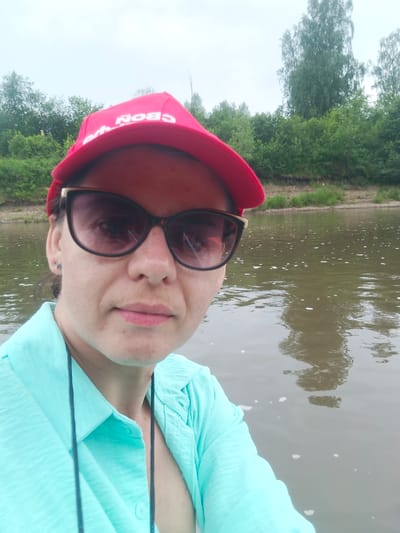 A woman wearing a red baseball cap and sunglasses is taking a selfie on a body of water. She has on a light blue collared shirt. The background consists of trees lining a riverbank. The scene appears to be in Yakhino, Russia, suggested by the presence of birch trees and the overcast sky, typical of the region.