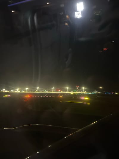 The image captures an aerial view at night, likely from an airplane window. The scene is dominated by the darkness of the night sky, punctuated by the lights of an airport or cityscape. In the foreground, a portion of an airplane wing is visible, its dark silhouette outlined by faint lights.
The middle ground displays a runway or tarmac area illuminated by numerous lights. These lights appear as streaks and blurs due to motion or atmospheric conditions, creating a vibrant and dynamic visual. Some lights are green, indicating direction, while others are red and yellow, possibly marking the edges of the runway. Tall lampposts with bright lights stand at intervals, casting cones of illumination onto the ground. In the distance, a cluster of buildings with illuminated windows and red roof lights suggests a built-up area.
The top portion of the image shows the interior of the aircraft cabin. Two illuminated signs are visible: a larger square sign with a bright white light, and a smaller rectangular sign below it, which appears to have white text on a dark background, possibly indicating an exit. Reflections on the window obscure some details of the cabin interior.
There is no specific information in the image to confirm the location as Moss Vale, Australia. The scene is generic to many airports at night.