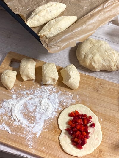 The image shows a top-down view of dough and chopped strawberries on a wooden cutting board and in a baking tray lined with parchment paper. The dough is divided into several portions, some of which are shaped into oval logs and placed in the baking tray, while others are in smaller, rounded pieces on the cutting board. One portion of dough on the cutting board is flattened and filled with chopped strawberries. There is also a large ball of dough in the background. The scene appears to be a kitchen, with a wooden surface visible beneath the cutting board. There are no people or animals visible in the image.