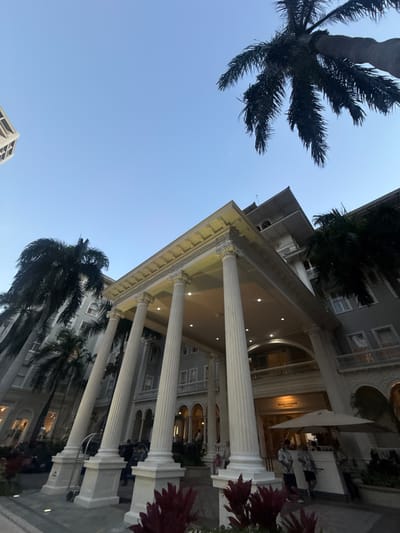 The image depicts the Moana Surfrider, A Westin Resort & Spa, Waikiki Beach, in Honolulu, United States of America, viewed from a low angle. Palm trees frame the sky and the building, with classical-style columns supporting an elaborate pediment. Some people are visible inside the building.