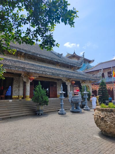 The image depicts the exterior of a Buddhist temple in Tuy An Nam, Vietnam, under a clear blue sky with scattered clouds. The temple features traditional East Asian architectural elements, including multi-tiered roofs with ornate eaves and decorative motifs. The building's facade is constructed of stone with intricate detailing. In front of the temple, a paved courtyard is visible, with stone steps leading up to the main entrance. Several large stone incense burners and decorative lanterns are placed in the courtyard. Two individuals are present: one man in a white t-shirt and shorts, and another man in a blue shirt and dark shorts. A flag with red and yellow colors is visible on a flagpole to the right of the temple. Lush green trees and potted plants are scattered around the temple grounds, with foliage from a large tree partially obscuring the top left of the frame. The overall atmosphere is serene and spiritual, characteristic of a religious site.