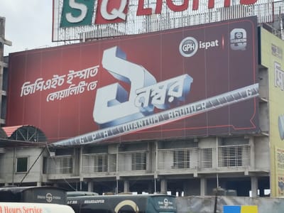 The image captures an urban scene, likely in Hatikumrul, Bangladesh, featuring a large billboard advertisement dominating the view. The advertisement is for "GPH Ispat" and "GPH Quantum B6000-R," showcasing a stylized representation of the number "1" in Bengali script.
Below the billboard is a building with multiple levels and barred windows. Several buses are parked in front of the building. Some buses have the word "Dhaka," followed by an image, and the word "Palma." Another bus has the words "A/C BUS SHAHZADPUR." The background includes overcast skies.