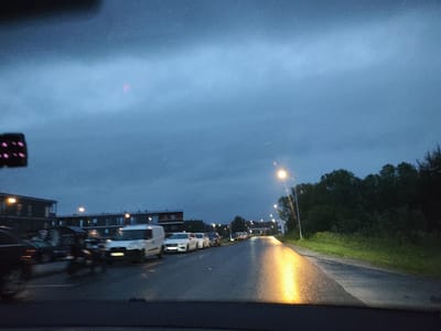 The image depicts a street scene in Tiskre, Estonia, during twilight or early evening. The sky is a deep blue-grey, with some lighter patches suggesting clouds. A street lamp on the right side of the road casts a strong, bright yellow reflection on the wet asphalt. Several cars are parked along the left side of the road. In the foreground, a white van is visible, along with other vehicles that are blurred due to motion or low light. Beyond the parked cars, apartment buildings are visible. Trees and greenery line the right side of the road. The overall impression is of a quiet, possibly residential, street at dusk.