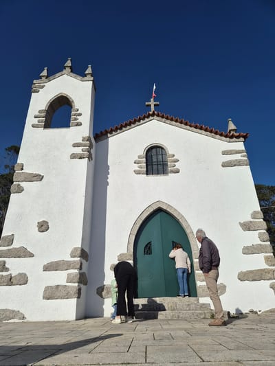 The image depicts the Igreja de São João Baptista, a church in Esposende, Portugal. The church is a white building with stone trim, a red-tiled roof, and a cross on top. Several people are standing in front of the church's green arched doors. One person, wearing khaki pants and a dark jacket, appears to be looking at the doors. Another person, wearing blue jeans and a white jacket, is also looking at the doors. A third person, wearing black pants and a green shirt, is also present. The sky is clear and blue. The church is located on a stone surface.