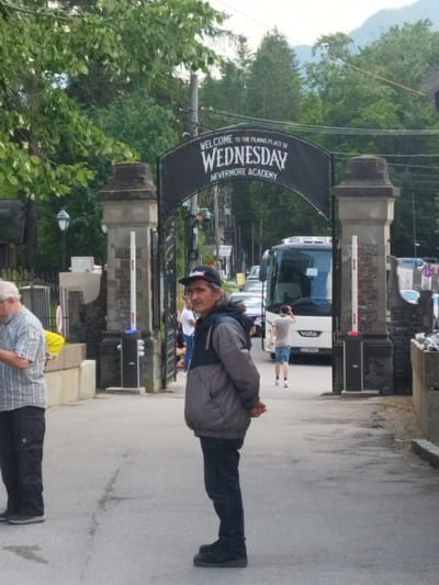 The image shows an outdoor scene in what appears to be an entryway to a facility, possibly an academy, indicated by a large banner overhead that reads "WELCOME TO THE FILMING PLACE OF WEDNESDAY NEVERMORE ACADEMY". Two men are visible in the foreground. The man on the left is wearing a striped shirt and dark pants, and he is looking down. The man on the right, closer to the camera, is wearing a dark jacket, dark pants, and a baseball cap. He is facing the camera. Behind him, a bus is partially visible, and other people are in the background. The environment includes stone pillars, a metal gate, and lush green trees. The scene is likely a public area leading to a filming location for the show "Wednesday" at Nevermore Academy, which is located in the city of Bușteni, Romania.