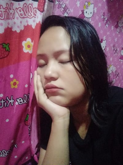 A young woman with dark hair rests her head on her hand, eyes closed. Her expression appears tired or bored. She wears a black top. The background consists of two pink, patterned textiles. One features Hello Kitty imagery, while the other has strawberries, flowers, and "Hello Kitty" lettering. The lighting is dim.