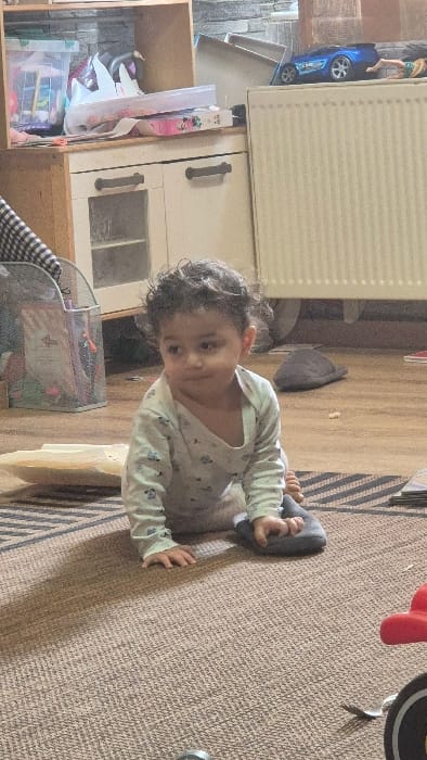 This image captures an indoor residential scene in Melle, Belgium. A single child, appearing to be a toddler with dark, curly hair and brown skin, is positioned on a light brown woven rug with darker striped patterns. The child wears a light-colored long-sleeve onesie patterned with small grey designs, possibly sheep or clouds. The child is crouched, resting on their knees and hands, facing left with a slight turn of the head and a subtle smile. A single dark grey slipper is worn on the child's right foot; the left foot is bare.
In the immediate foreground to the right, a portion of a red and black toy, resembling a tricycle or ride-on toy, is visible. To the left of the child, a piece of light-colored, possibly paper or thin cardboard, lies flat on the rug.
The background features a light-colored wooden floor extending behind the rug. Against the back wall, a multi-component storage unit is present. On the left, a light wooden shelving unit houses clear plastic bins containing various items, alongside papers and bags. Below this, a white cabinet with two doors and dark horizontal handles is visible, with its upper cabinet window revealing interior shelving. To the right of the cabinet, a tall, vertical white radiator is mounted against the wall. A blue toy car and a partially visible doll (resembling Barbie) are positioned on top of the radiator. The wall behind the radiator and cabinet appears to have a textured grey, possibly stone-like, finish. A metal mesh bin containing items is visible to the left, behind the child, adjacent to what appears to be a checkered fabric item. The scene is well-lit, casting soft shadows.