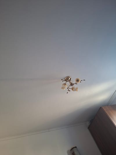 An indoor ceiling with a light fixture is shown. The light fixture is made of gold-colored metal and has three tulip-shaped glass shades. A dark brown cabinet is visible in the bottom right corner of the image, along with part of a rolled-up rug. The ceiling is white and there is a thin horizontal line across it. The location is the city of Zaşağan, Kazakhstan.
