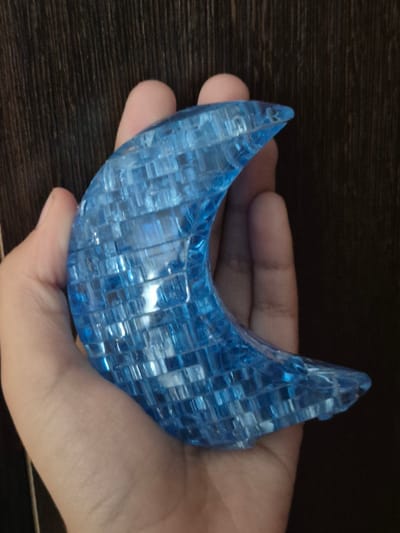 A close-up shot shows a translucent blue crescent-shaped puzzle piece being held by a person's hand against a dark, vertically-grained wooden background. The puzzle piece is made up of many small, interlocking rectangular segments. The hand is cupped, supporting the puzzle piece from below and the sides. The lighting highlights the reflective and transparent qualities of the puzzle. The image is an uncropped, full-view photograph. There is no information available regarding the location in the city of Новоишимское, Kazakhstan.