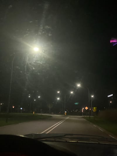 The image depicts a night view of a street in Waalwijk, Netherlands. Streetlights line the left side of the road, illuminating the area. The road itself has white lane markings and appears wet, possibly from recent rain. A vehicle interior is visible on the right side, suggesting the photo was taken from inside a car. In the distance, traffic signs and other street furniture can be discerned. In the upper left, there is a purple light.