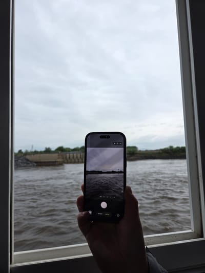 The image presents a view through a white window frame, looking out at a wide body of water under an overcast sky. In the bottom right, a person's hand is visible, holding a dark smartphone that is actively displaying its camera interface.

The scene outside the window is dominated by a vast expanse of choppy, muddy-brown water, which appears turbulent, suggesting strong currents or wind. In the distance, a blurry shoreline is visible, characterized by a line of green trees and what appear to be linear, possibly concrete or stone structures along the bank, which could be part of an embankment or dam infrastructure. The sky above is uniformly light grey, indicating a heavily overcast or cloudy day.

The smartphone, which appears to be an iPhone based on its design and camera app interface, is held vertically. Its screen shows a live preview of a scene closely resembling the outdoor view, with the water, distant land, and sky displayed horizontally within the phone's frame. The camera application's controls are visible on the screen, featuring Cyrillic text where "ФОТО" (Photo) is highlighted, next to "ВИДЕО" (Video). Additionally, zoom options such as "0.5x", "1x", "2x", "4x", and "8x" are present along the right edge of the screen. The person is engaged in photographing or videoing the river and landscape.

The overall lighting is diffused and dim, consistent with an overcast daytime. Given the location context of Nizhneleninskoe, Russia, this scene likely captures a significant river, such as the Amur, which forms part of the region's landscape.