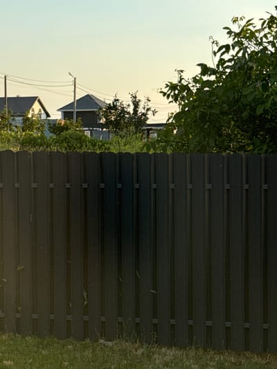 The image depicts a residential scene in Tsibanobalka, Russia. The primary focus is a tall, dark, corrugated metal fence that dominates the right side of the frame. To the left of the fence, a residential area is visible under a pale, hazy sky, suggesting it might be early morning or late afternoon, or perhaps an overcast day.

In the background, the rooftops of several houses are discernible, characterized by dark, angled shingles. Some of these houses have visible windows and architectural details. Electrical poles with wires stretch across the sky, indicating the presence of utilities. Lush green foliage from trees and bushes partially obscures the houses, adding a natural element to the scene. The grass in the foreground, to the right of the fence, is a vibrant green.

The composition, with the fence prominently featured, creates a sense of division or enclosure. The overall mood appears calm and quiet, without any human presence or overt activity. The natural light suggests a peaceful, everyday setting. No visible text is present in the image.
