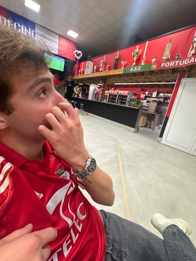 An indoor scene within a Portuguese community establishment, likely a bar or club, in Décines-Charpieu, France, is captured. The foreground features a male individual, seen from the right side of his head and upper torso, wearing a red short-sleeved S.L. Benfica football jersey and dark grey jeans. He wears a metallic watch on his left wrist and has a small tattoo on his left forearm. His right hand is raised to his face, fingers near his nose, as he gazes upwards and to the left towards a television screen displaying a green football pitch.
The background reveals a red wall adorned with a wooden shelf displaying multiple sports trophies, including golden cups and statuettes. A prominent banner on this wall reads "A.S.C PORTUGAL" with a crest featuring Portuguese flag colors. Two decorative heart-shaped objects are also on the shelf. Above this, a banner on the left side reads "DÉCINOIS" adjacent to vertical French flag colors. Below the shelf, a long, dark-colored bar counter extends across the scene. Behind this counter, one male individual in a green shirt is visible. The ceiling is dark with recessed lighting, and the floor is a light-colored concrete with a yellow line marking.