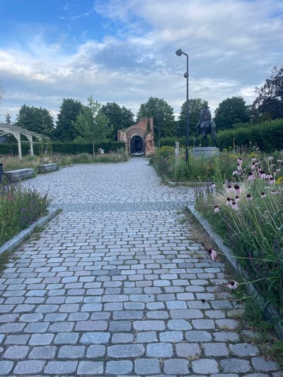 This is a landscape photograph of a cobblestone path leading towards an arched brick structure in Weert, Netherlands. The path is lined with greenery and flowering plants, including coneflowers, which are mostly in bloom. To the left of the path, there is a statue of a horse and rider. In the background, trees form a dense green backdrop, and above them, an overcast sky with patches of blue is visible. The image appears to be taken during the daytime. The scene evokes a sense of tranquility and historical ambiance, with the cobblestone path and brick arch suggesting an older area. No visible text is present in the image.
