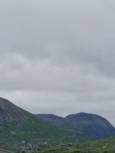 The image depicts a mountainous landscape with a village nestled at the base of the mountains. The sky is overcast with clouds. The village appears to consist of scattered buildings with dark roofs, surrounded by green vegetation. The mountains are rocky and covered with patches of green foliage. The scene suggests a rural setting in a mountainous region, potentially a settlement in Hovden, Norway, given the context. There are no people or animals visible in the image, nor any apparent actions or interactions. The overall impression is one of a quiet, natural environment.
