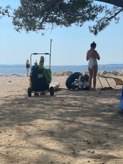 This image depicts a beach scene in Crikvenica, Croatia, with the Adriatic Sea visible in the background. In the foreground, sand is scattered with pine needles and small pebbles. Two women are present. One woman, wearing a white swimsuit, stands facing away from the camera. To her left, a large black wagon is loaded with beach equipment, including a life ring, towels, and a surfboard. Another woman, wearing a black swimsuit and a white hat, stands to the left of the wagon, looking towards the sea. A beach chair and other assorted items are scattered on the sand. The scene is framed by the branches of a pine tree overhead.