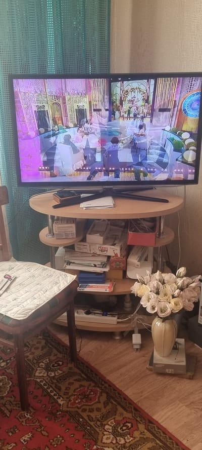 The media file shows an indoor scene with a television displaying an animated image of people in a brightly lit, ornate setting. A wooden table with rounded edges stands beside the television, holding various objects including books, boxes, and a power strip. A bouquet of light-colored flowers sits on top of the table, next to a decorative vase. Further back, a chair with a patterned cushion is visible, and a portion of a red carpet with intricate designs covers the floor. The lighting suggests it is daytime. Some visible text on boxes includes "ГЛАВ ТОРТ" and "ВЕНОЛТОН 911". The overall impression is of a lived-in domestic space, possibly a living room or study. The location context is Krymsk, Russia.