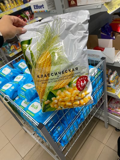 A person's hand with light-colored nail polish is holding a large, clear plastic bag of corn snacks. The bag features an illustration of a corn cob and kernels, along with Russian text that reads "КЛАССИЧЕСКАЯ КУКУРУЗНАЯ ПАЛОЧКА В САХАРНОЙ ПУДРЕ ВКУС ДЕТСТВА ДЛЯ ВСЕЙ СЕМЬИ" and a red "FAMILY PACK" logo with figures of a family. The bag is positioned in front of a shopping cart filled with blue packages of "Eco Life" wet wipes. The shopping cart is in a store aisle, with shelves of products visible in the background. A sign reading "Effort" is visible on an upper shelf. The flooring appears to be tiled. The image was taken in Kushnarenkovo, Russia.