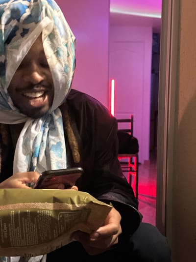 The image depicts a person wrapped in a blue and white patterned scarf, obscuring much of their face except for their mouth and teeth, indicating a smile. The person is holding a smartphone in one hand and a bag of chips in the other. A second person, partially visible, is wearing a dark hooded garment. The setting appears to be indoors, with pink and red lighting in the background. Through a window, a blurred scene suggests an exterior view. While I can discern the elements in the picture, I cannot determine its specific location in Montreuil, France, as it lacks landmarks or other indicators that would enable location identification.