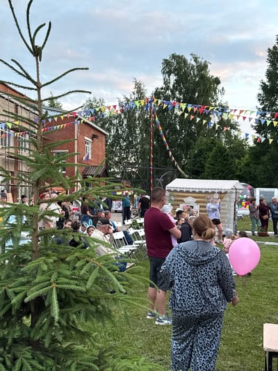 The image depicts an outdoor gathering in a village setting, possibly in the city of Kukuí, Russia, given the context provided. In the foreground, a large evergreen tree dominates the left side of the frame. Behind the tree, a crowd of people are assembled on a grassy area. Several individuals are seated on white folding chairs, facing a small, wooden structure decorated with what appears to be a painted facade resembling a traditional Russian izba.
A woman in a leopard-print tracksuit is walking away from the camera towards the right, holding a large pink balloon. A man in a burgundy t-shirt is holding a baby. In the background, a brick building with scaffolding is visible. Colorful triangular flags are strung across the scene, strung between poles and trees, suggesting a festive or celebratory atmosphere. The sky is overcast with some hints of color, indicating it might be late afternoon or early evening. Various trees and foliage are scattered throughout the background.