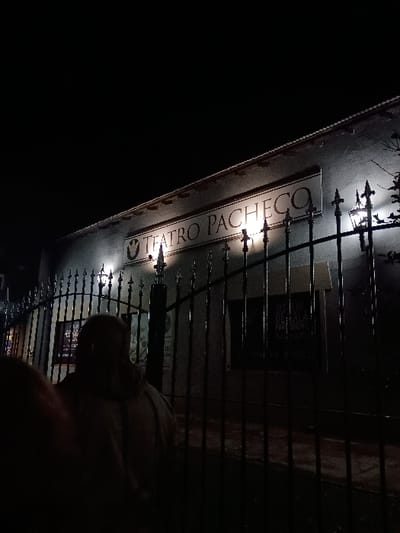 The image shows a night-time view of the Teatro Pacheco in General Pacheco, Argentina, located at the coordinates -34.457 latitude and -58.633 longitude. The building, partially obscured by a black wrought iron gate, has a large sign reading "TEATRO PACHECO" above a window. The sign is illuminated by a nearby streetlight. A single person is visible, standing behind the gate, obscured by darkness. The image suggests an urban, nighttime setting with a focus on the theater building. The person's presence suggests potential activity or interaction with the theater. The overall image is dark and somewhat mysterious.