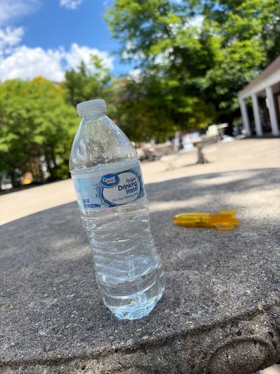 The image captures an outdoor scene, likely in Edison, United States of America, under bright daylight conditions. The main subjects are a plastic water bottle and a small, yellow, bomb-shaped object, both resting on a textured, light grey concrete or asphalt surface.
The "Great Value Purified Drinking Water" bottle, appearing partially full and adorned with water droplets, lies horizontally on its side. Its label is clearly visible, reading: "Great Value Purified Drinking Water With Flavor-Enhancing Minerals 16.9 FL OZ (1PT 0.9 FL OZ) 500mL". Adjacent to it, further into the frame, is a distinct yellow object resembling a stylized bomb or a toy, with fins at one end.
The background, largely out of focus, reveals vibrant green foliage from trees and a clear blue sky dotted with scattered white clouds, indicating a sunny and pleasant day. Shadows cast on the ground suggest the sun is fairly high in the sky, perhaps midday or early afternoon. No specific activity is taking place; the image presents a static arrangement of these items in an outdoor setting, possibly a park, pavement, or a common area next to a building, as implied by blurred structures in the far background.