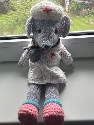 A crocheted stuffed mouse wearing a nurse's hat and uniform sits on a windowsill. The mouse is gray with black button eyes and pink crocheted feet with light blue cuffs. Its uniform consists of a white coat with red cross emblems and a small gray pouch attached to the side. The nurse's hat is white with a red cross. The background is a slightly out-of-focus view of greenery seen through a window with a screen, suggesting an outdoor environment. The windowsill is white.