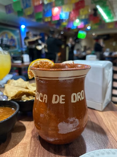A cantaro cup with "Boca de Oro" on it rests on a wooden table in Guadalajara, Mexico. The cup is brown with white stripes and lettering. It holds a drink, garnished with a slice of orange. Next to it is a white dispenser, chips, and a bowl of salsa. The background shows a restaurant scene with colorful decorations and blurred figures of people.