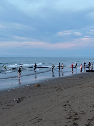 A group of fishermen are pulling a large fishing net from the ocean onto a sandy beach in Uleekareung, Indonesia. The sky is overcast with hues of blue and pink, indicating either dawn or dusk. The water is calm with gentle waves lapping at the shore. The fishermen, dressed in various casual clothing, are positioned along the net, some in the shallow water and others on the wet sand, all appearing to be engaged in the strenuous task of hauling the net. Several individuals are clearly visible, with their postures suggesting exertion. On the beach, there are tracks from vehicles or possibly a boat, and a coiled rope lies on the sand. A discarded plastic bottle is also visible on the beach. The overall scene depicts a communal fishing effort in a coastal Indonesian setting.