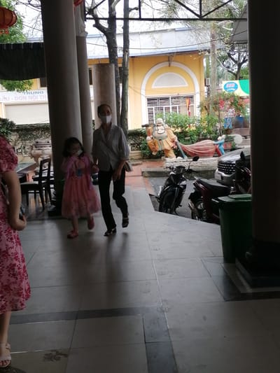 Captured from an indoor perspective in Chu Văn An Ward, Vietnam, the image shows a woman and a young girl walking towards the camera. Both are wearing face masks. The woman is in a button-down shirt and dark pants, while the girl wears a pink dress. A figure in a floral dress is partially visible in the lower left corner. Outside, various scooters and cars are visible, along with statues of a Buddha and other figures. A yellow building with arched windows stands in the background. The shot is framed by pillars, suggesting the view is taken from a covered area.